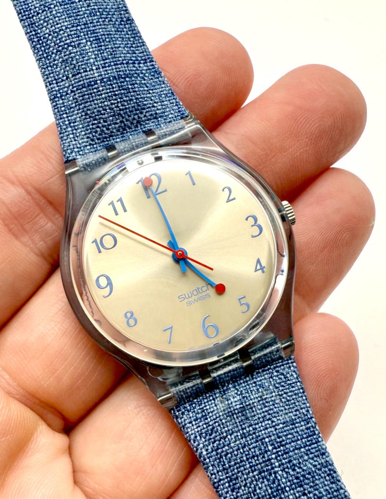 May include: A Swatch Swiss watch with a light-coloured face, blue numerals, and blue and red hands. The watch has a clear plastic case and a blue denim-style strap.