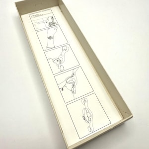 May include: An open, rectangular box with a light beige exterior. Inside, a series of black and white line drawings illustrate a process, likely for a product. The drawings are on a cream-colored paper.