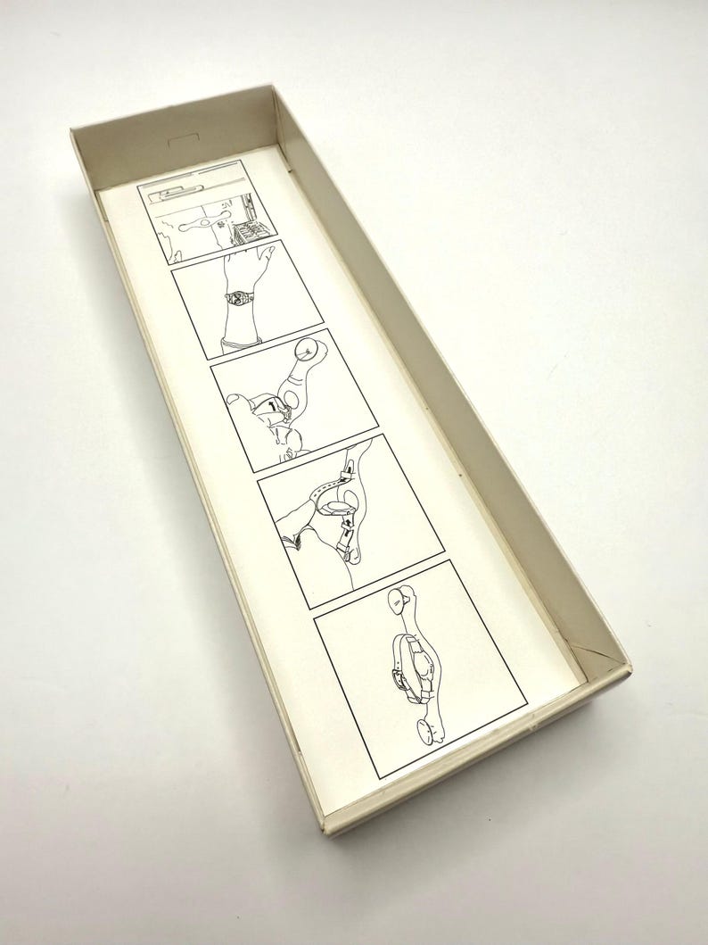 May include: An open, rectangular box with a light beige exterior. Inside, a series of black and white line drawings illustrate a process, likely for a product. The drawings are on a cream-colored paper.