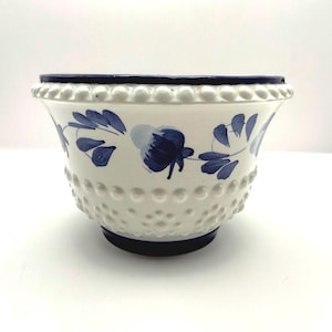 May include: A white ceramic bowl featuring a raised hobnail texture on the exterior and a hand-painted blue floral pattern encircling the middle. The bowl has a dark blue rim and a dark blue base, with a wide, flared opening at the top.