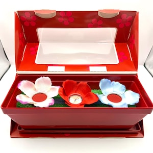 May include: A red box with a clear lid, containing three flower-shaped watches. The watches are in shades of pink, red, and blue, resting on green faux grass. The box has a floral design.