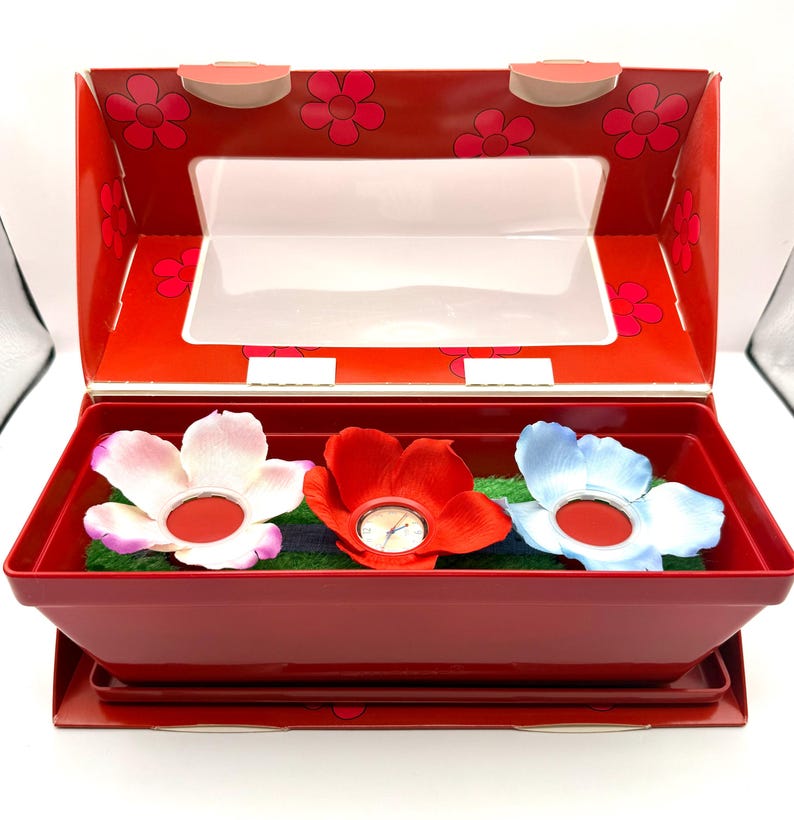 May include: A red box with a clear lid, containing three flower-shaped watches. The watches are in shades of pink, red, and blue, resting on green faux grass. The box has a floral design.