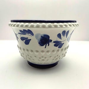 May include: A white ceramic bowl featuring a raised hobnail texture across the exterior surface. The rim and base are accented with a dark blue band. A hand-painted blue floral pattern with leaves encircles the upper portion of the bowl.