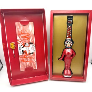 May include: A red Swatch watch in its original box. The watch features a red and black strap with a repeating pattern. The watch face is a woman's face. The box is red with a gold interior.