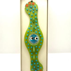 May include: A unique green watch with a whimsical design, featuring a round blue watch face with cartoon eyes. The watch band is shaped like a stylized foot with blue and orange accents. The watch is presented in a white box.
