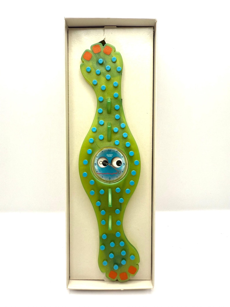 May include: A unique green watch with a whimsical design, featuring a round blue watch face with cartoon eyes. The watch band is shaped like a stylized foot with blue and orange accents. The watch is presented in a white box.