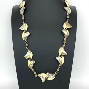 May include: A silver-toned necklace with a series of leaf-shaped pendants. Each pendant has a cream-colored center and a curved, silver-colored edge. The necklace is displayed on a black mannequin.