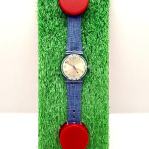 May include: A wristwatch with a denim blue strap and a clear plastic case. The watch face is gold with black numerals. Two red circular accents are attached to the strap. The watch is displayed on a green, textured surface.