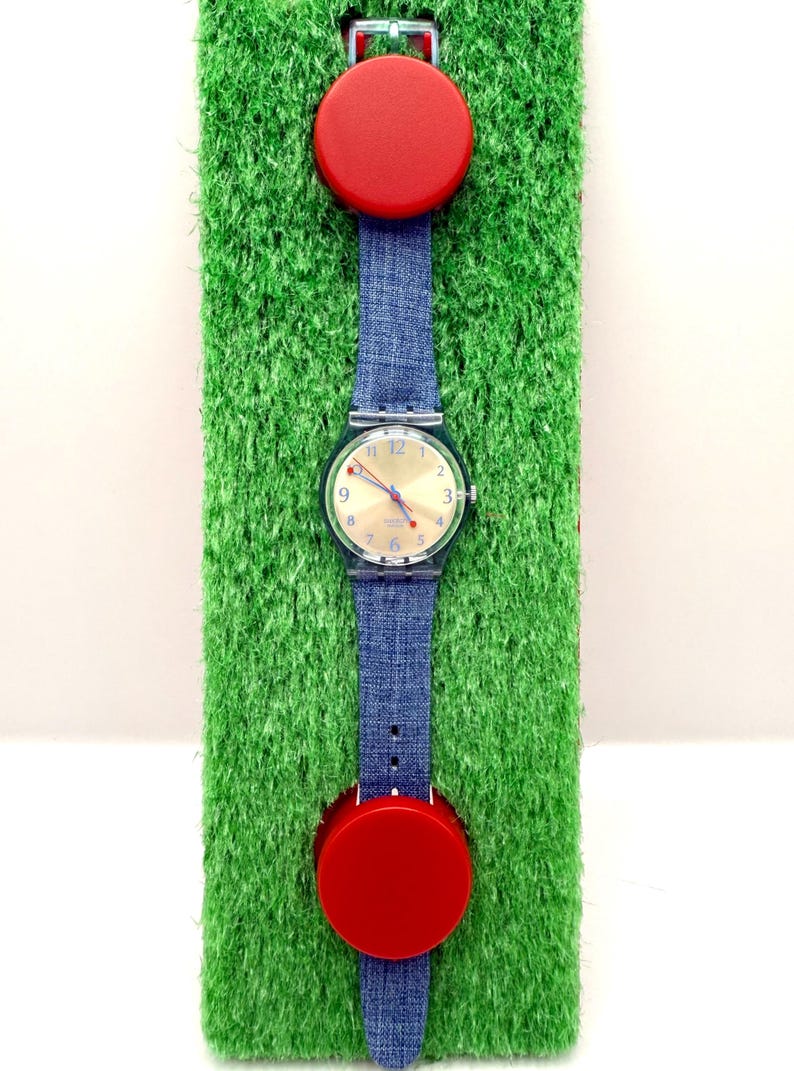 May include: A wristwatch with a denim blue strap and a clear plastic case. The watch face is gold with black numerals. Two red circular accents are attached to the strap. The watch is displayed on a green, textured surface.