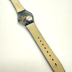 May include: A clear Swatch watch with a transparent case, revealing the internal workings. It has a light-colored leather strap with blue denim detailing and a clear buckle. The watch strap is approximately 24 cm in length.