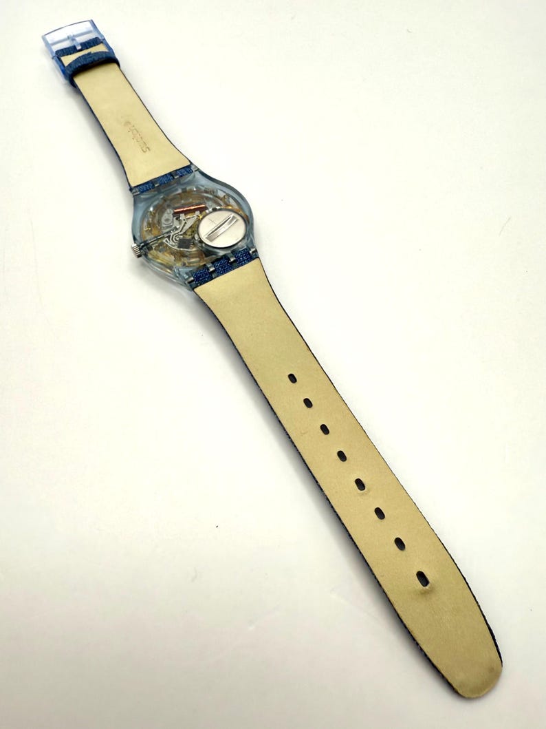 May include: A clear Swatch watch with a transparent case, revealing the internal workings. It has a light-colored leather strap with blue denim detailing and a clear buckle. The watch strap is approximately 24 cm in length.