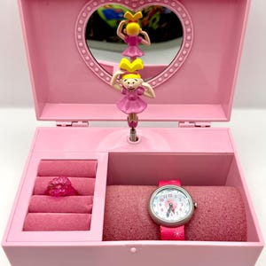 May include: Pink musical jewelry box with a heart-shaped mirror and ballerina figurines. The box contains a ring and a watch with a pink band. The watch face has a floral design.