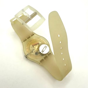 May include: A transparent, light yellow wristwatch with a matching strap and buckle. The watch face reveals the internal mechanisms. The strap has multiple holes for adjustment. The watch is lying on a white surface.
