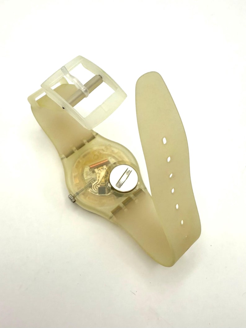 May include: A transparent, light yellow wristwatch with a matching strap and buckle. The watch face reveals the internal mechanisms. The strap has multiple holes for adjustment. The watch is lying on a white surface.