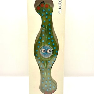 May include: A Swatch watch in its original packaging. The watch has a green, organic shape with blue polka dots and red accents. The watch face features a whimsical design. The packaging is a cream color with the word "Swatch" printed on the side.