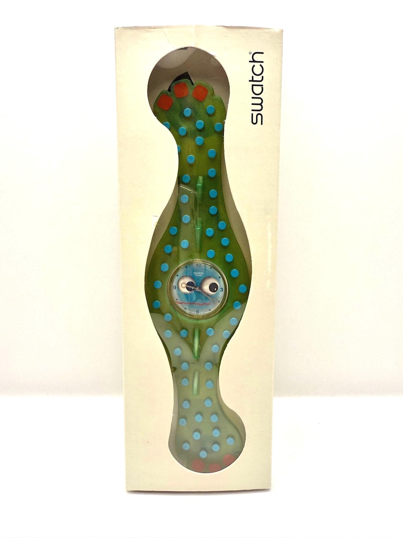 May include: A Swatch watch in its original packaging. The watch has a green, organic shape with blue polka dots and red accents. The watch face features a whimsical design. The packaging is a cream color with the word "Swatch" printed on the side.