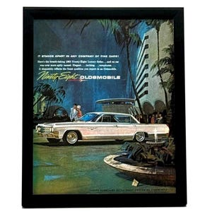 May include: A framed vintage advertisement for a 1963 Oldsmobile Ninety-Eight Luxury Sedan. The print features a white car parked in front of a modern building with palm trees and a fountain, including the text: IT STANDS APART IN ANY COMPANY OF FINE CARS!