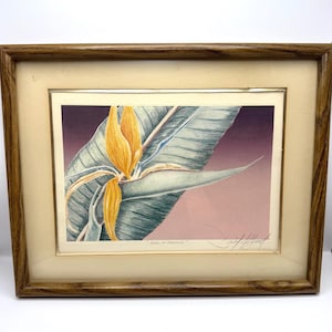 May include: A framed print titled Bird of Paradise featuring a yellow flower and large green leaves against a purple and pink gradient background. The artwork is enclosed in a light-colored mat and a medium-toned wooden frame with a visible signature.