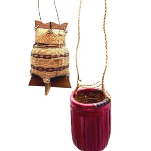 May include: A woven lidded basket with a natural tan finish, red and black geometric accents, and a wooden base, paired with a cylindrical woven basket dyed in a vibrant magenta pink, both featuring thin cord handles for hanging or carrying.