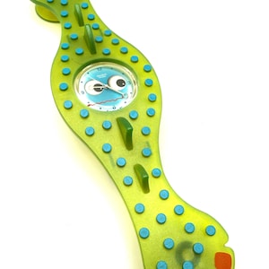May include: A whimsical, green wristwatch with a cartoonish face on the watch face. The watch has a unique, elongated shape with blue and orange accents. The watch is a fun, playful accessory.