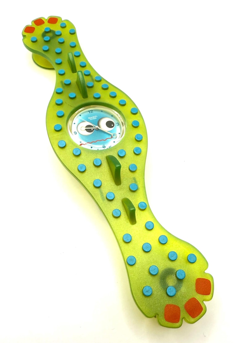 May include: A whimsical, green wristwatch with a cartoonish face on the watch face. The watch has a unique, elongated shape with blue and orange accents. The watch is a fun, playful accessory.