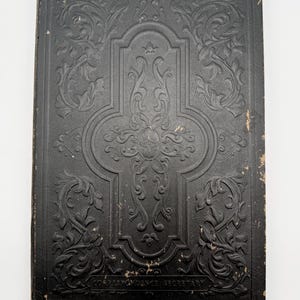 May include: A black rectangular book with an embossed cover featuring intricate scrollwork and floral patterns. The center displays a cross-shaped design, and the text CORRESPONDENCE SECRETARY is printed in small capital letters at the bottom.
