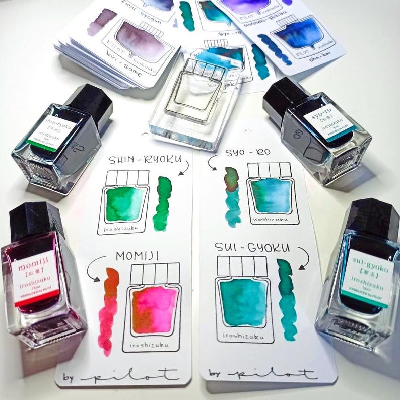 May include: An assortment of Pilot Iroshizuku ink bottles with black caps and labels, alongside color swatches. The inks are in various colors, including green, blue, and red. The image includes the text "Iroshizuku" and "by pilot".