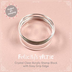 May include: A clear acrylic stamp block with an easy grip edge. The stamp block is round and has a transparent appearance. The text "Felicity's Attic" and "Crystal Clear Acrylic Stamp Block with Easy Grip Edge" are visible.