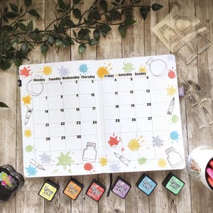 our premium quality stamps can be used for creative journaling and monthly planner pages, the image features a monthly planner page with colourful ink splatters and ink bottles. Distress Ink pads and art supplies are arranged around the planner on a wooden surface.