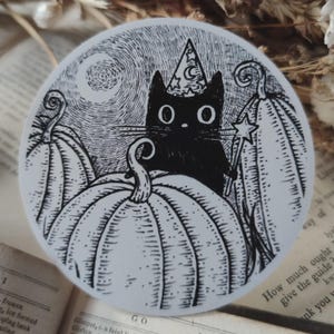 May include: A circular sticker featuring a black and white ink illustration of a black cat wearing a star and moon patterned wizard hat. The cat holds a star-tipped wand and is surrounded by three large pumpkins under a swirling night sky.