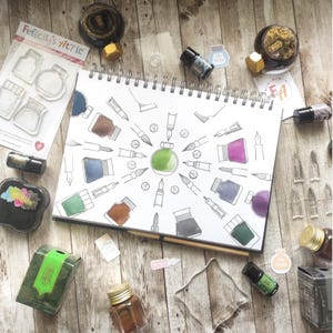 May include: A collection of art supplies, including ink bottles, stamps, and a sketchbook with pen and ink illustrations. The sketchbook features a central green ink bottle design surrounded by various pen and ink drawings. The text "Felicity's Attic" is visible.