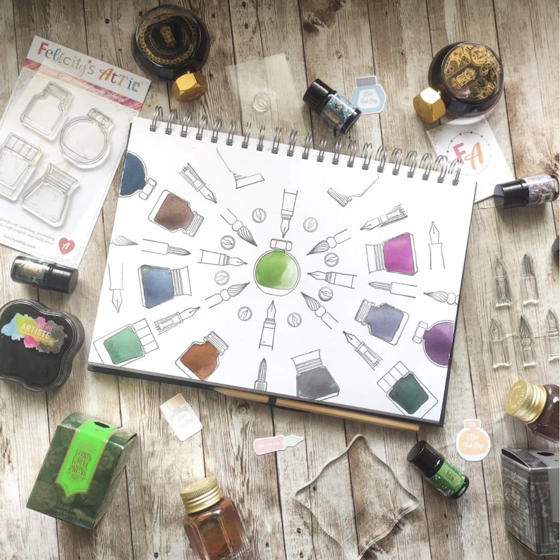 May include: A collection of art supplies, including ink bottles, stamps, and a sketchbook with pen and ink illustrations. The sketchbook features a central green ink bottle design surrounded by various pen and ink drawings. The text "Felicity's Attic" is visible.