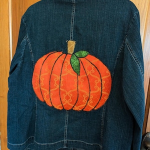 May include: A dark blue denim jacket with a large orange pumpkin applique on the back. The pumpkin has a green stem and leaves.