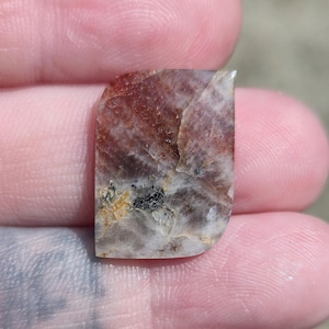 May include: A polished, rectangular gemstone with a rounded top and a flat bottom. The stone features a mix of colours, including reddish-brown, white, and black, with a marbled appearance. The gemstone is held between fingers.