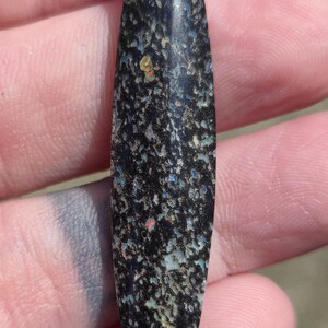 May include: A polished, elongated gemstone with a dark, speckled base color. The stone features a mosaic of iridescent colors, including blue, green, and red, creating a unique visual texture. The shape is smooth and symmetrical.