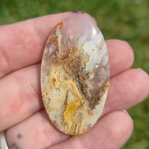 Puede incluir: Un cabujón de piedra preciosa pulida de forma ovalada con una base rosa pálido translúcida e inclusiones dendríticas ramificadas complejas en tonos amarillo dorado, marrón oscuro y blanco crema, creando un patrón natural, orgánico y estratificado.