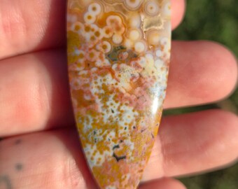 Op de afbeelding: Een gepolijste, druppelvormige edelsteen met een uniek patroon. De steen heeft een mix van kleuren, waaronder oranje, geel, wit en vleugjes blauw en zwart. Het oppervlak heeft een glanzende afwerking, die de natuurlijke variaties benadrukt.