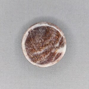May include: A circular decorative item with a marbled effect. The centre displays a blend of brown and white tones with gold specks, enclosed by a white edge. The object has a polished, smooth surface.