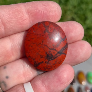 May include: A round, polished gemstone with a deep red base color and intricate black veining patterns. The stone is held in a hand, showcasing its smooth, domed surface and natural variations. The gemstone is likely used for jewelry or decorative purposes.