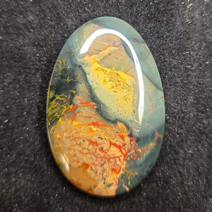 May include: A polished oval-shaped gemstone with a mix of green, black, yellow, and red colors. The stone has a unique pattern of veins and swirls.
