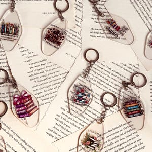 May include: Transparent acrylic keyrings featuring book-related designs, such as stacks of books, floral details, and phrases like "Booktrovert" and "Just one more chapter." Each keyring has a silver ring.