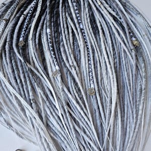 Puede incluir: Una colección de rastas grises y blancas, con diferentes tonos y texturas. Algunas rastas están adornadas con abalorios plateados, incluyendo estrellas y espirales. El efecto general es una mezcla de tonos claros y oscuros, creando un peinado visualmente interesante.