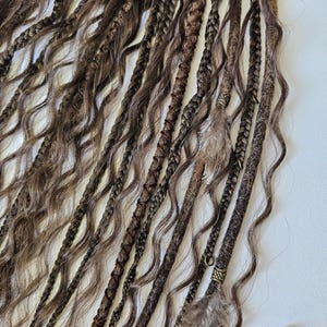 May include: A set of brown and blonde synthetic dreadlocks with brown leather and feather accents. The dreadlocks are arranged in a row and have a wavy texture.