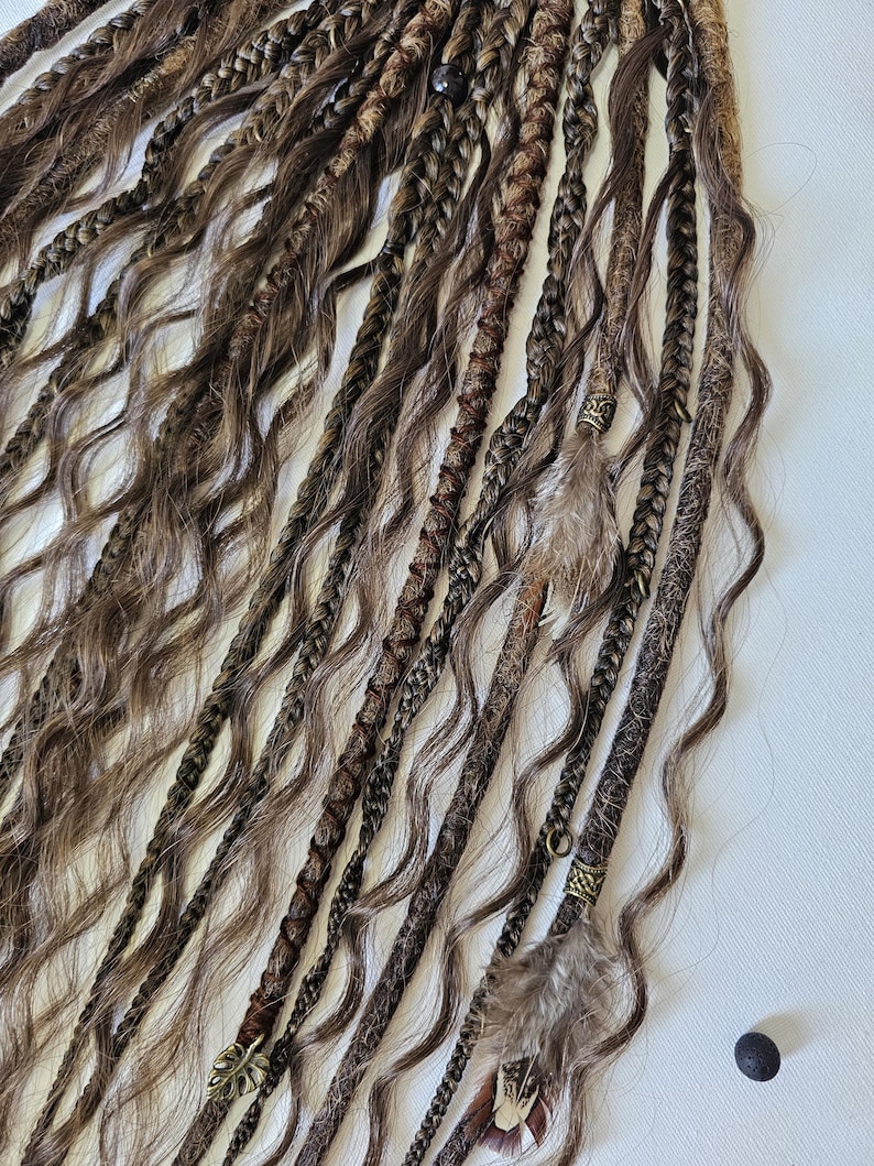 May include: A set of brown and blonde synthetic dreadlocks with brown leather and feather accents. The dreadlocks are arranged in a row and have a wavy texture.