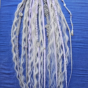 Può includere: Una collezione di dreadlock fatti a mano nei toni del grigio, lavanda e bianco. I dread presentano varie trame, tra cui sezioni lisce, intrecciate e ondulate. Ciondoli e perline d'argento sono intervallati tra i fili, aggiungendo dettagli decorativi. Lo sfondo è un tessuto blu.