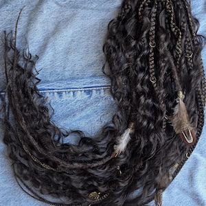 Op de afbeelding: Donkerbruine krullende hairextensions met een zwarte haarband. De extensions zijn versierd met kleine goudkleurige metalen ringen, veren en decoratieve bedels. De hairextensions zijn ontworpen om als paardenstaart te worden gedragen.