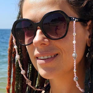 以下が含まれることがあります： ピンクのビーズの眼鏡チェーンをかけた黒いサングラスをかけた女性。チェーンには銀と灰色のビーズと手の形をした小さなチャームが付いています。