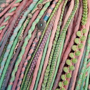 Puede incluir: Primer plano de un conjunto de rastas sintéticas en tonos rosa, verde y azul. Las rastas están decoradas con cuentas y abalorios.