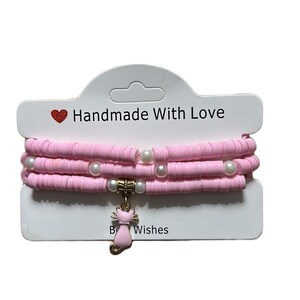 May include: A set of pink beaded bracelets with white pearl accents and a gold-coloured cat charm. The card reads "Handmade With Love" and "Best Wishes". The bracelets are displayed on a white card.