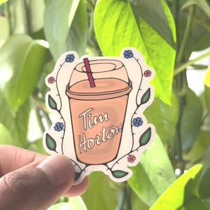 May include: A sticker of a Tim Hortons coffee cup with a straw, surrounded by a floral design with blue and pink flowers. The cup is beige with the text "Tim Hortons" in brown.